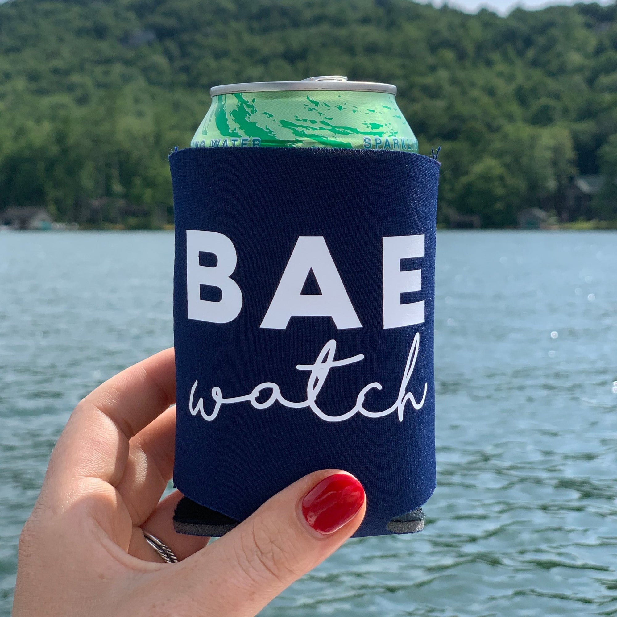 A navy can cooler is held up which reads "BAE watch" with a backdrop of a lake.