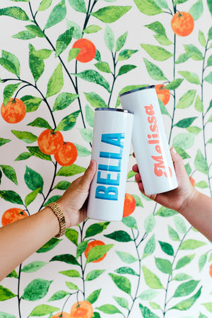 Two people hold up their custom white tumblers in front of a wall painted with oranges and leaves.