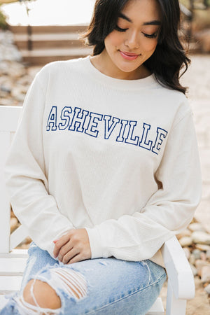 A girl sits on a bench showing off her white corded sweatshirt which reads "Asheville" in navy.