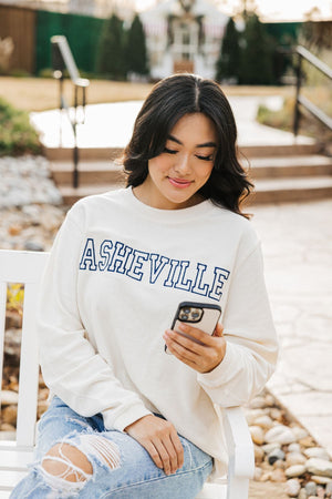 A girl sits on a bench looking at her phone and wearing a white corded sweatshirt which reads "Asheville" in navy.