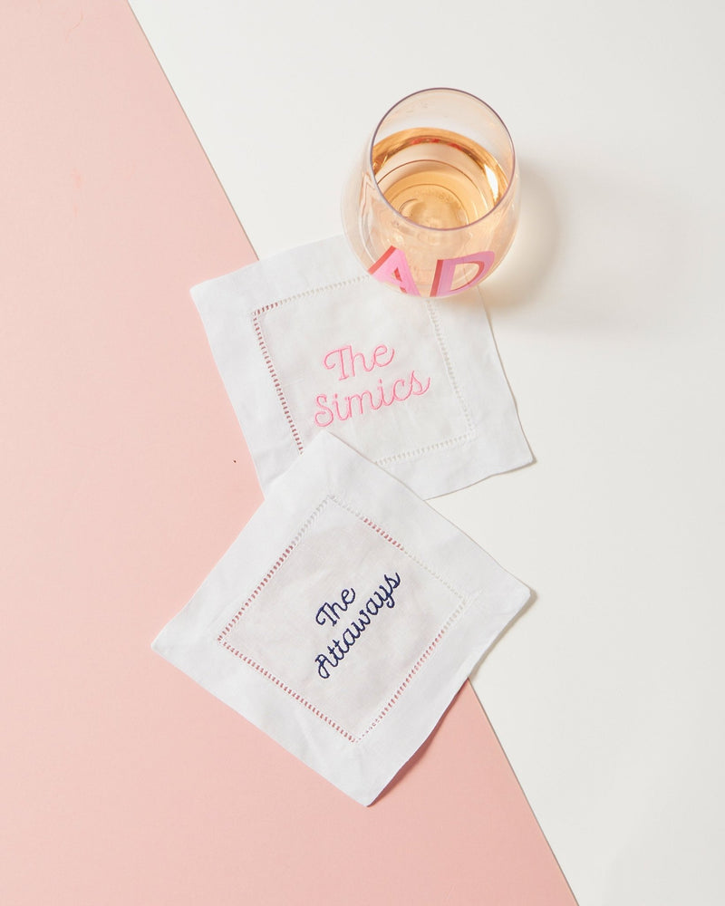Two cocktail napkins are set out with a wine glass to show off some of the embroidered names that can customize your cocktail napkins.