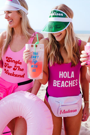 Two girls on the beach wear pink custom swimsuits, fanny packs and visors. 