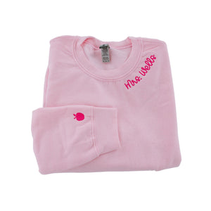 Folded pink crewneck sweatshirt with 'Mrs. Wells' embroidered in pink script on the collar and a small pink apple icon embroidered on the cuff