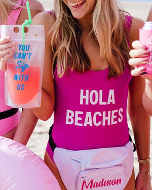 A fuchsia swimsuit is customized with "Hola Beaches" written in white text