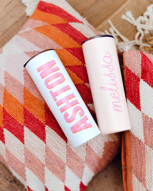 Two custom white tumblers with different lettering options sit on a pillow