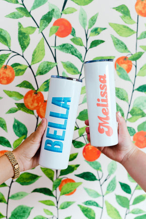 Two people hold up their custom white tumblers in front of a wall painted with oranges and leaves.