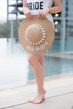 A woman at a pool holds her custom floppy beach hat which reads "Mrs. Dillon" in white.