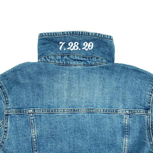 A jean jacket is laid down with the collar up showing a custom date printed to it.