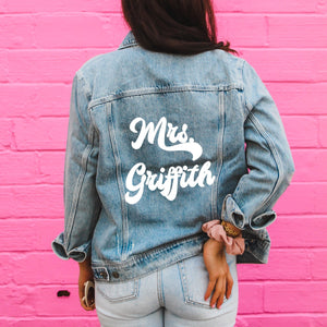 A woman in front of a pink wall wears a jacket which reads "Mrs. Griffith" across the back.