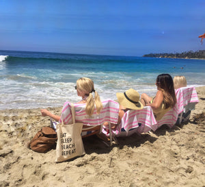 A group of women sit at the beach with a "Eat Sleep Beach Repeat" tote hanging on one of their chairs.