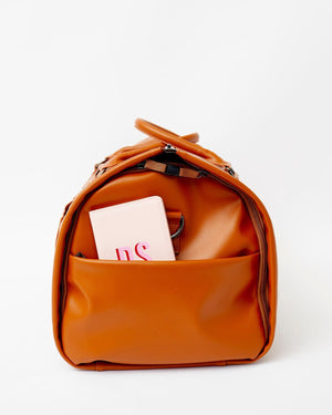 A white passport holder is placed into the side pocket of a tan duffel bag.