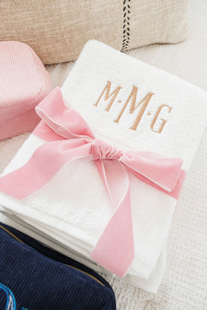 A white towel is embroidered with a beige monogram and is wrapped in a velvet pink bow