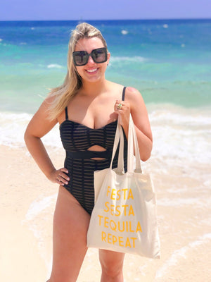 A woman stands on the beach holding her tote which says "Fiesta Siesta Tequila Repeat" in yellow.