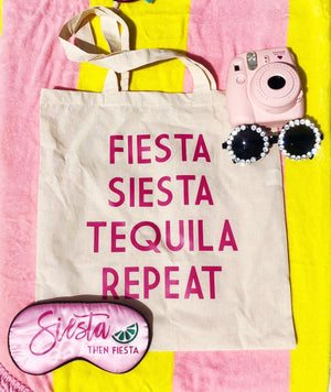 A tote with "Fiesta Siesta Tequila Repeat" printed in pink sits with a custom sleep mask and a pair of sunglasses.