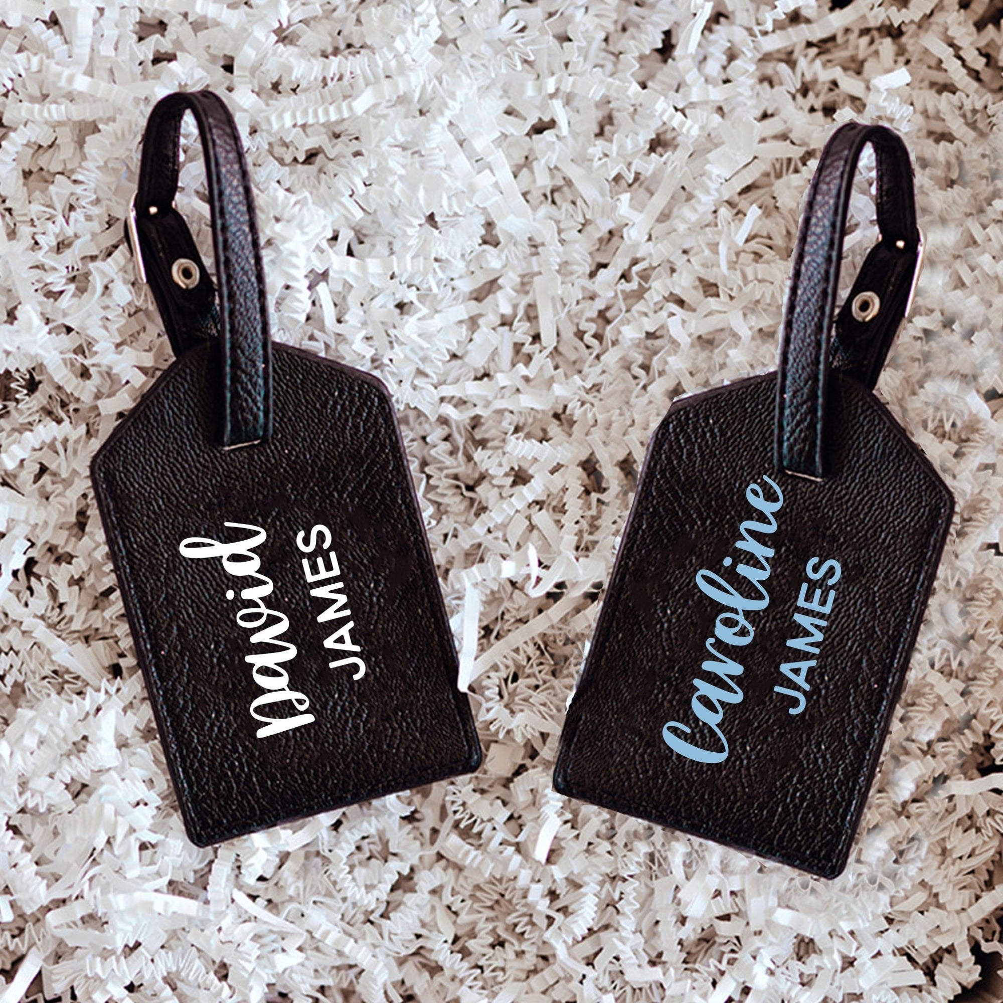 Two black luggage tags are personalized with names in a white and a blue font