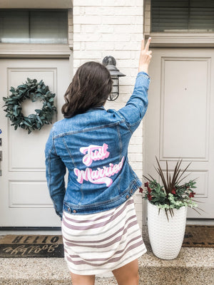 A woman stands outside showing off her custom denim jacket which reads "Just Married" in pink and white.