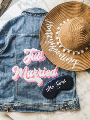 An assortment of products is laid out for the bride including a jean jacket which reads "Just Married," a sleep mask that reads "Mrs. Simic," and a floppy beach hat which reads "honeymooning."