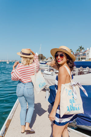Two women stand holding totes that read "let's get nauti" in bright blue