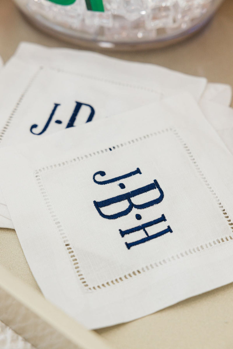 A set of cocktail napkins is embroidered with a monogram in a navy thread.