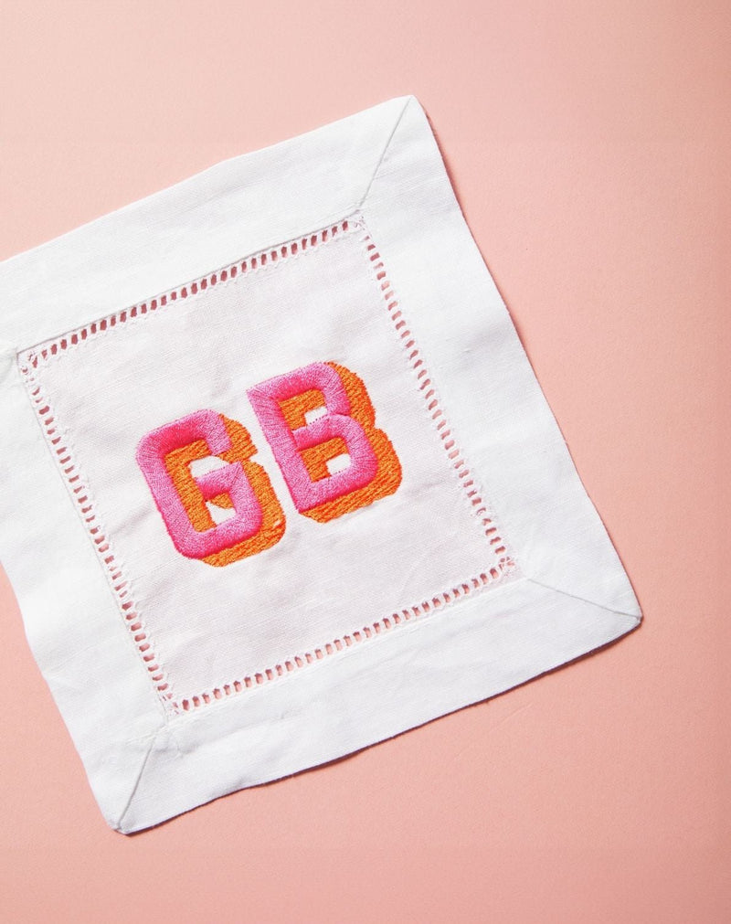 A cocktail napkin is customized with an embroidered block shadow monogram in pink and orange thread colors.