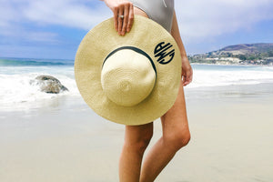A woman on the beach is holding a floppy beach hat with a black circular monogram.