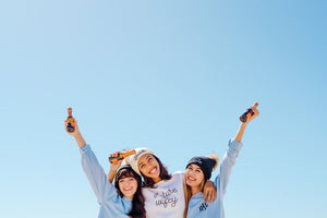 A group of women stand together in sweatshirts and beanies holding up mini champagne bottles.