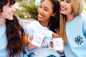 A group of women in white and blue sweatshirts cheers with their custom mugs.