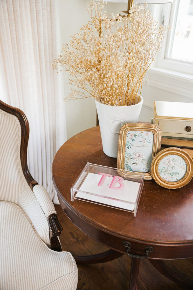 A pink monogrammed acrylic catchall box is used to collect cards on a brown wooden table.