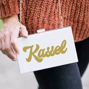A white clutch is customized with a name in gold font.