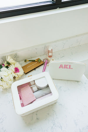 A white leather pouch is personalized with a pink monogram and a white leather clear case is personalized with a gold foil monogram.