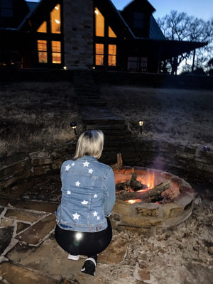 A woman sits in front of a fire pit wearing a customized jean jacket with white stars.