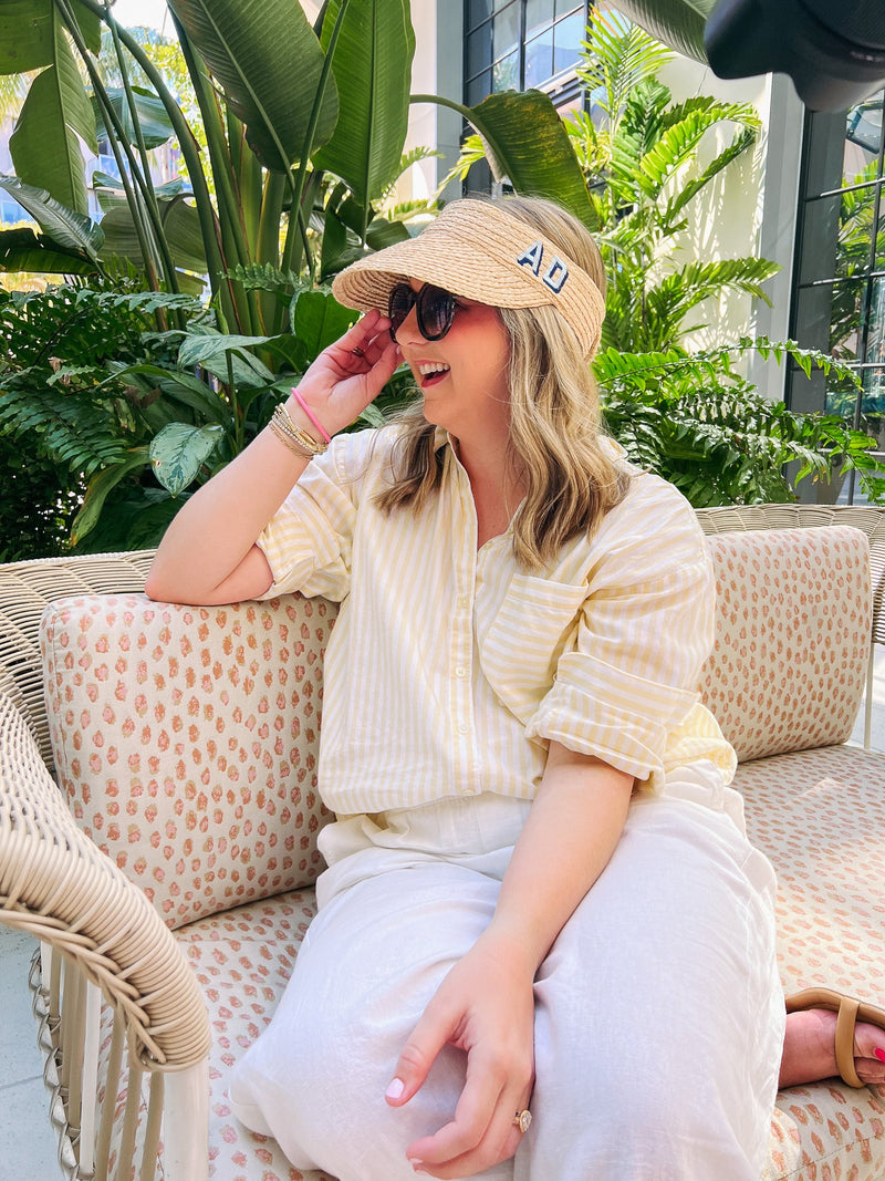 A girl sits on a couch by some plants wearing a straw visor with a blue monogram embroidered on it.