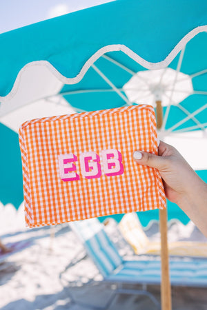 A person holds up an orange pouch with a pink monogram in front of a blue umbrella.