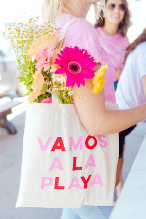 A tote bag filled with flowers is customized with the phrase "Vamos A La Playa" in pink and red.