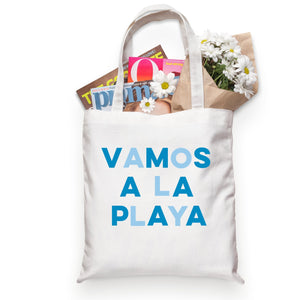 A white cotton tote reads "VAMOS A LA PLAYA" across the front in blue letters