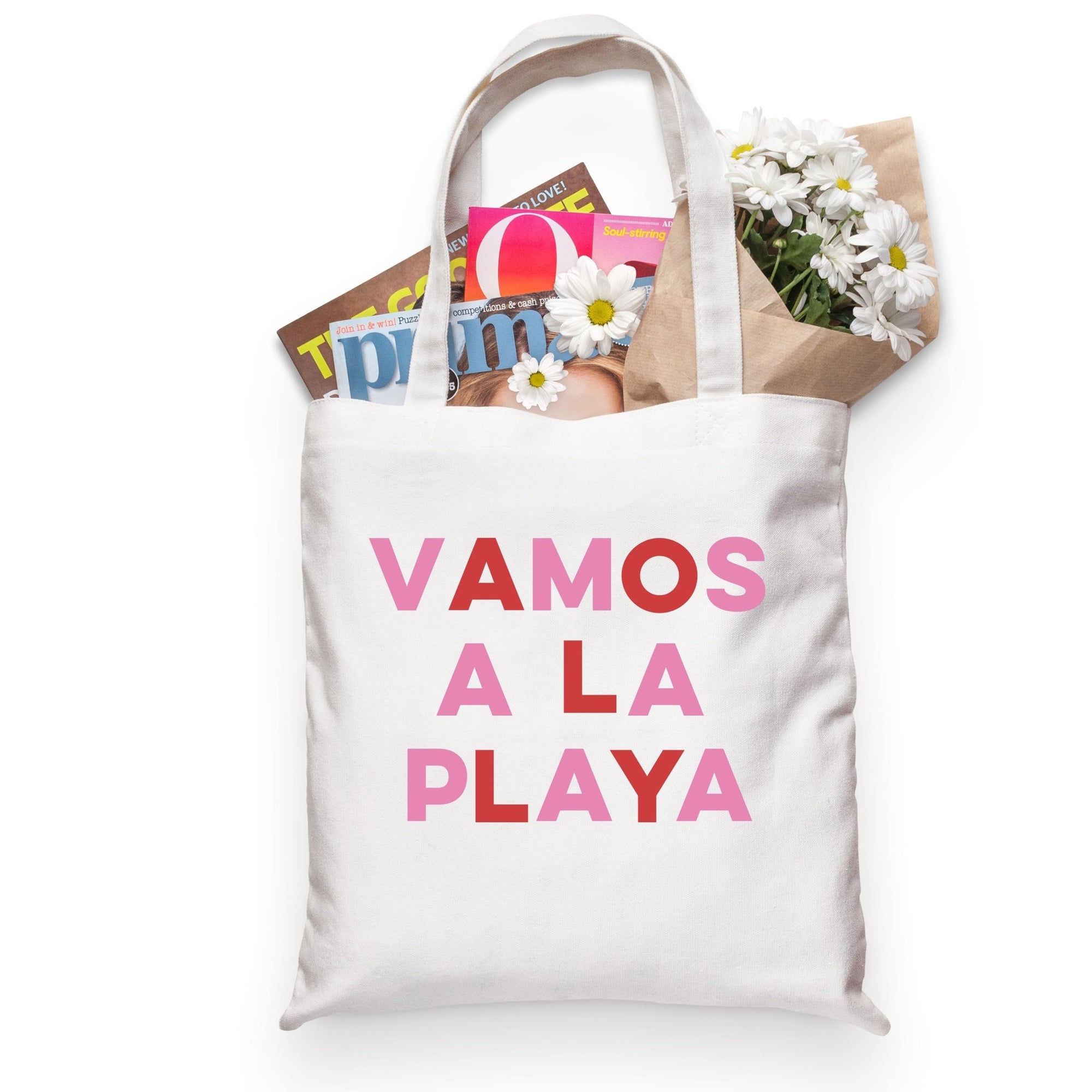 A tote bag filled with magazines and flowers is customized with the phrase "Vamos A La Playa" in pink and red.