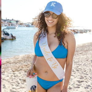 A woman on the beach wears a custom sash that says "Bride To Be" and a blue baseball hat with an initial on it