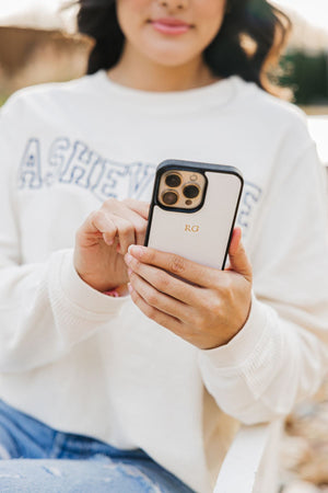 A woman in a white sweatshirt holds her phone which is in a case with a gold foil monogram