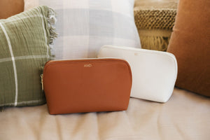 A tan and a white leather pouch are personalized with a gold foil monogram