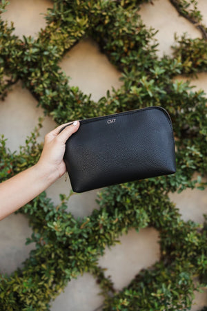 A hand holds a custom black vegan pouch with gold foil lettering next to a garden wall