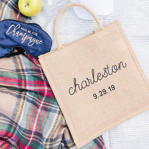 A jute tote bag is customized with a city and a date in black font.