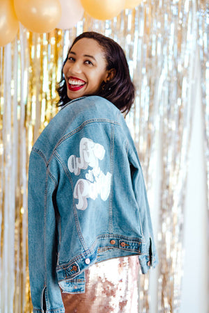 A woman holds a denim jacket that says "Wife of the Party" over her shoulder. 