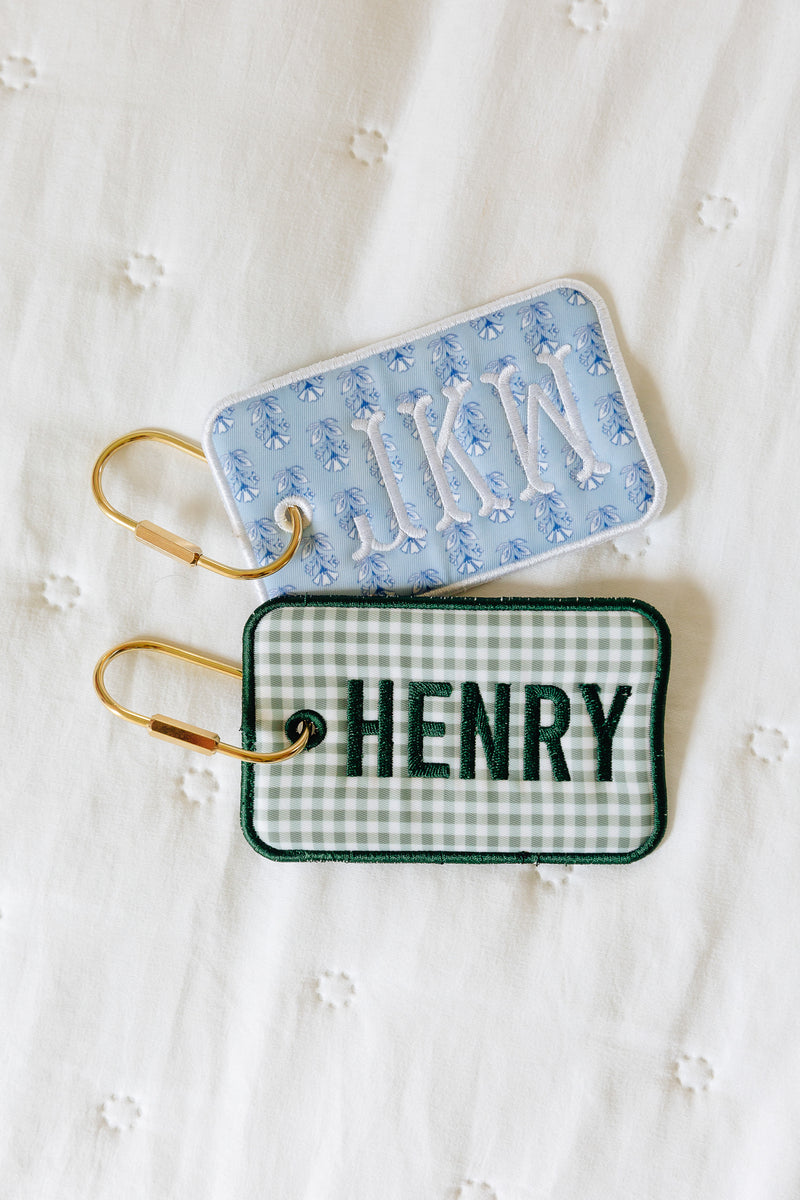 Two personalized embroidered luggage tags on gold key rings, displayed on a white embroidered fabric background. One tag has a light blue and white gingham pattern with white embroidered initials 'JKN'.