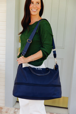 A woman wearing a green top and white pants carries a navy blue shoulder bag with a monogrammed 'CHS' on the front. The bag has a wide shoulder strap and a double-strap design, shown in front of a white door.