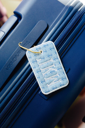 A light blue quilted luggage tag with white embroidered lettering and a gold metal ring, attached to a textured navy blue suitcase.
