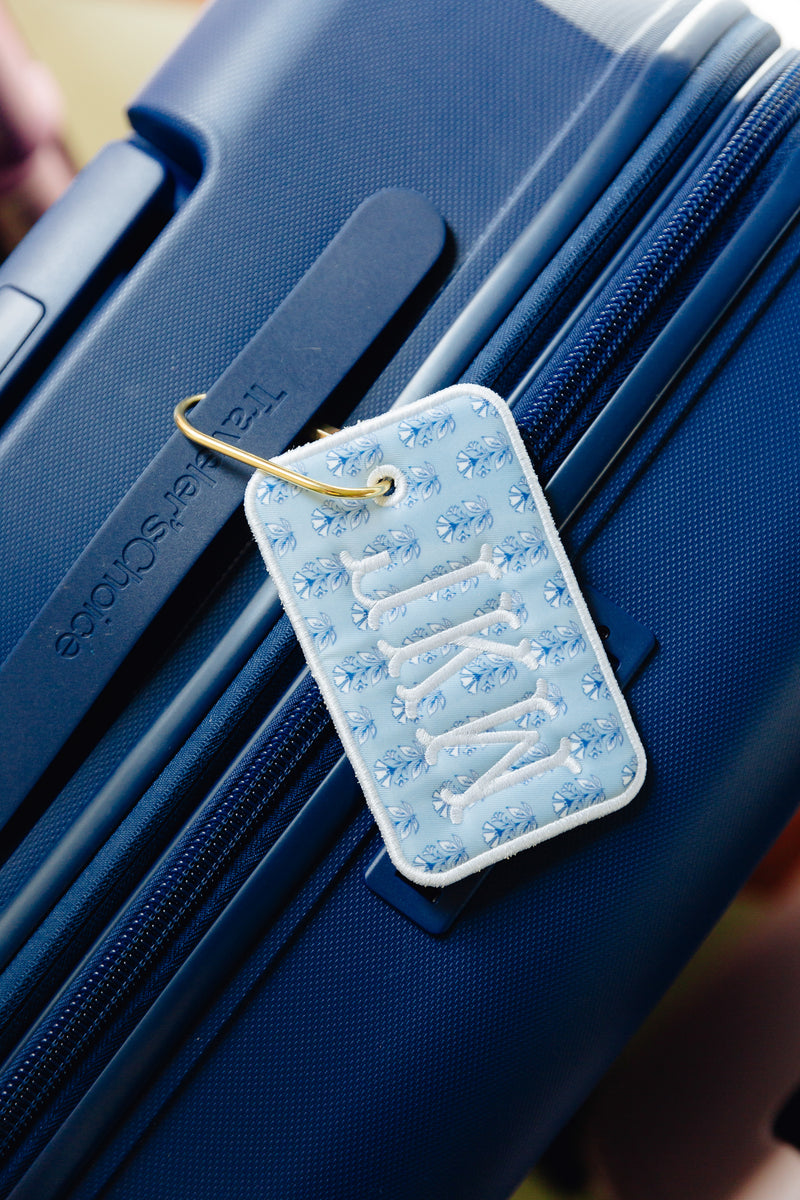 A light blue quilted luggage tag with white embroidered lettering and a gold metal ring, attached to a textured navy blue suitcase.