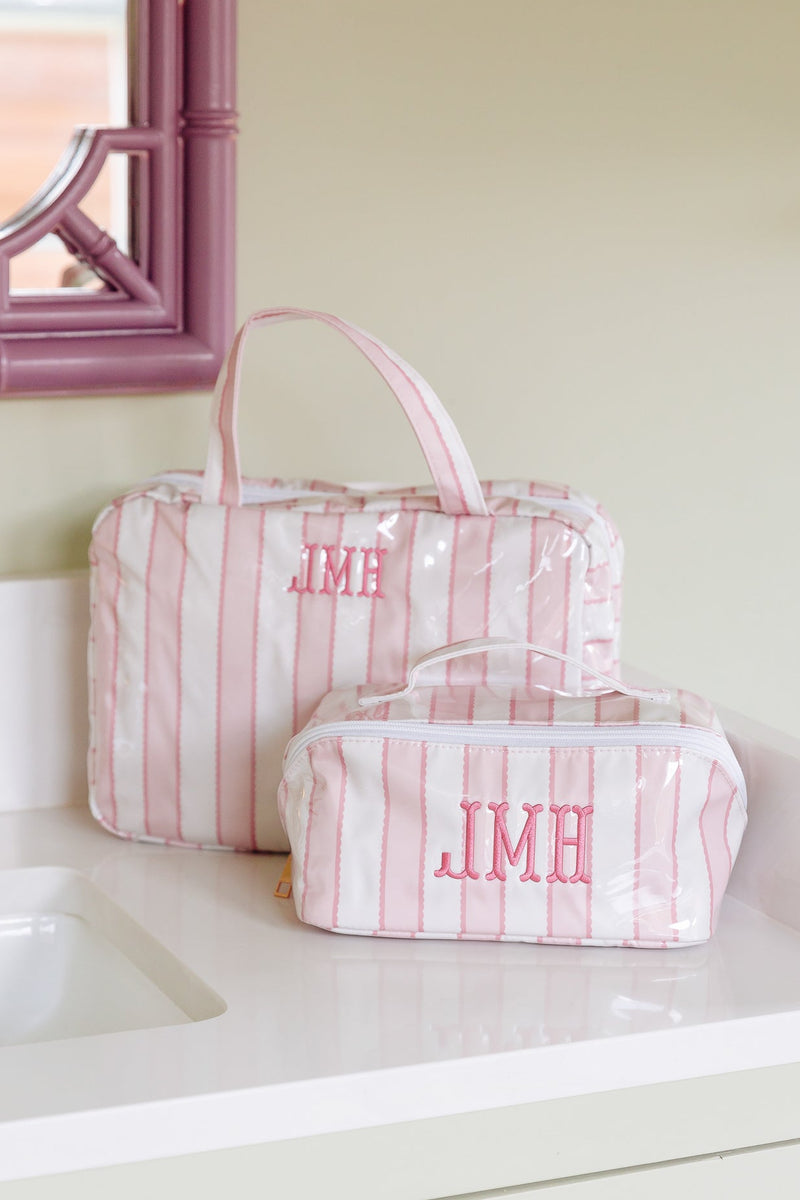 Two pink and white striped toiletry bags with pink monogrammed initials 'JMH' sit on a white bathroom counter next to a sink and a pink-framed mirror.