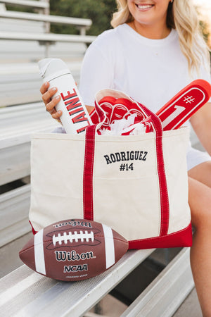 White 24 oz flip-lid tumbler monogrammed “LAINE” in red, held above a personalized gameday canvas tote