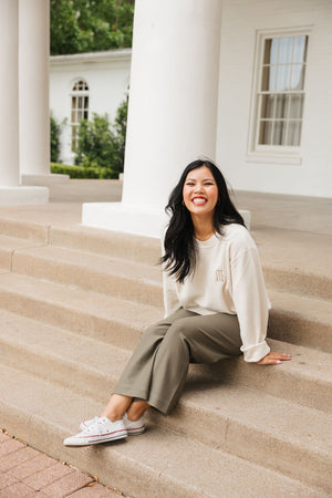 A woman sits on the steps smiling in her custom monogram corded sweatshirt.