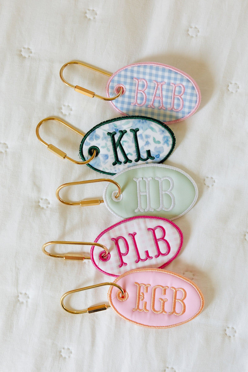 Five personalized oval keychains with gold carabiners are arranged vertically on a white textured fabric background. Each keychain has a different pastel color and monogram: pink gingham with 'BAB', blue floral with 'KL', pale green with 'HB', pink with 'PLB', and light pink with 'EGB'.